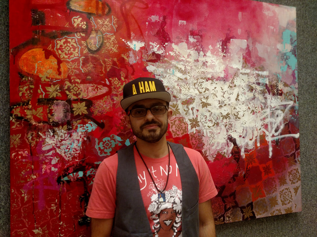 T.J. stands in front of a red and patterned painting. He is wearing a black rimmed hat with 